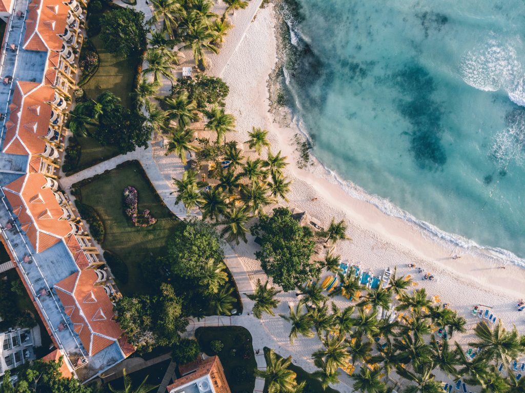 Aerial view of a tropical beach with turquoise water, palm trees, sandy shore, and a resort with orange rooftops. Lounge chairs are arranged along the beach near the water’s edge.