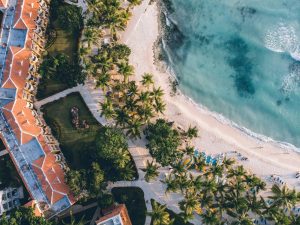 Aerial view of a tropical beach with turquoise water, palm trees, sandy shore, and a resort with orange rooftops. Lounge chairs are arranged along the beach near the water’s edge.