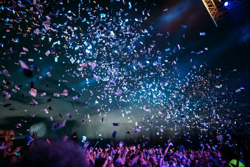 A large crowd at a concert or festival with colorful confetti and tickets raining down from above, illuminated by stage lights against a dark sky.
