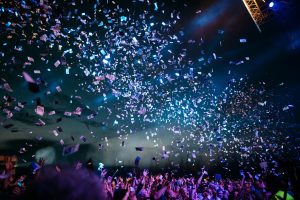 A large crowd at a concert or festival with colorful confetti and tickets raining down from above, illuminated by stage lights against a dark sky.