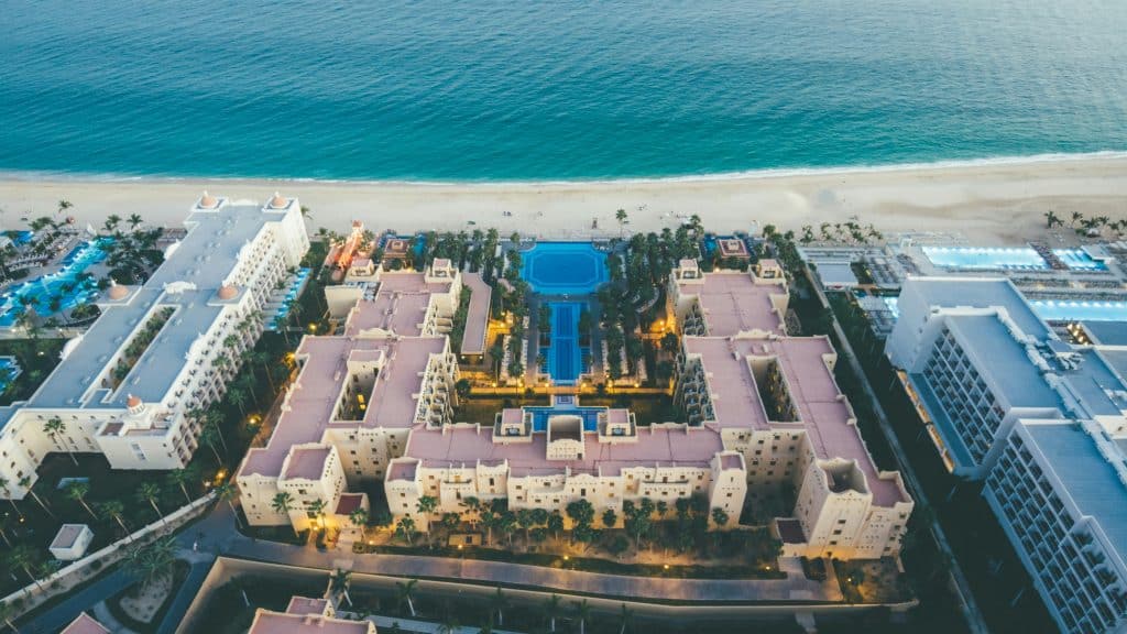 Aerial view of a large beachfront resort with multiple buildings, a central pool, palm trees, and lights, adjacent to sandy beach and blue ocean.