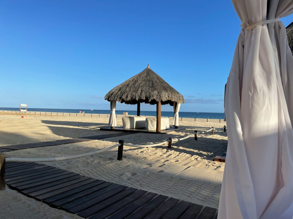A thatched-roof cabana with white lounge beds sits on a sandy beach near the ocean, with a wooden boardwalk and white curtains in the foreground under a clear blue sky.