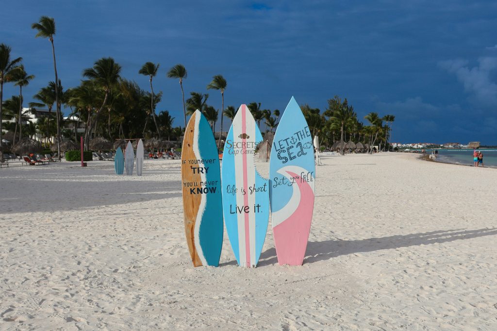Four colorful surfboards with inspirational quotes stand upright in the sand on a tropical beach lined with palm trees, with blue sky and ocean in the background.