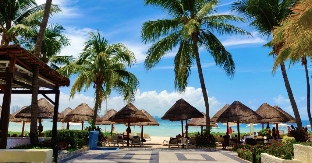 A tropical beach scene with tall palm trees, thatched umbrellas, lounge chairs, and people relaxing on the sand under a bright blue sky with scattered clouds. The ocean is visible in the background.
