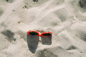 Red sunglasses with white arms lying on light-colored sand, casting small shadows.