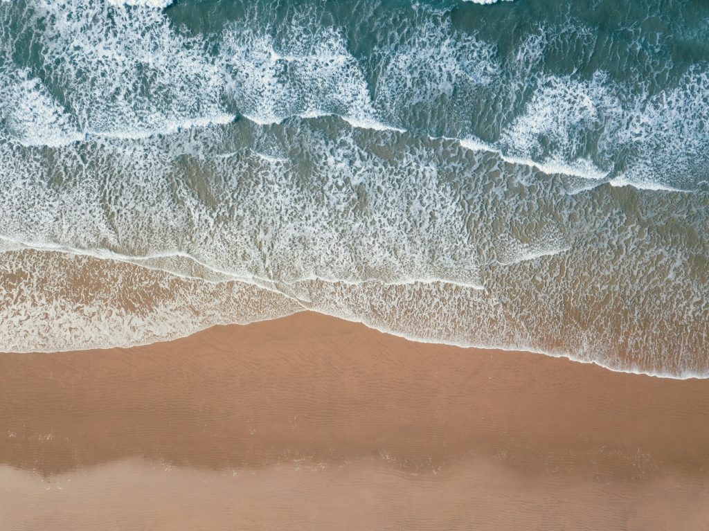 Aerial view of ocean waves gently rolling onto a sandy beach, with white foam forming patterns along the shoreline. The water transitions from deep blue-green to light brown near the sand.