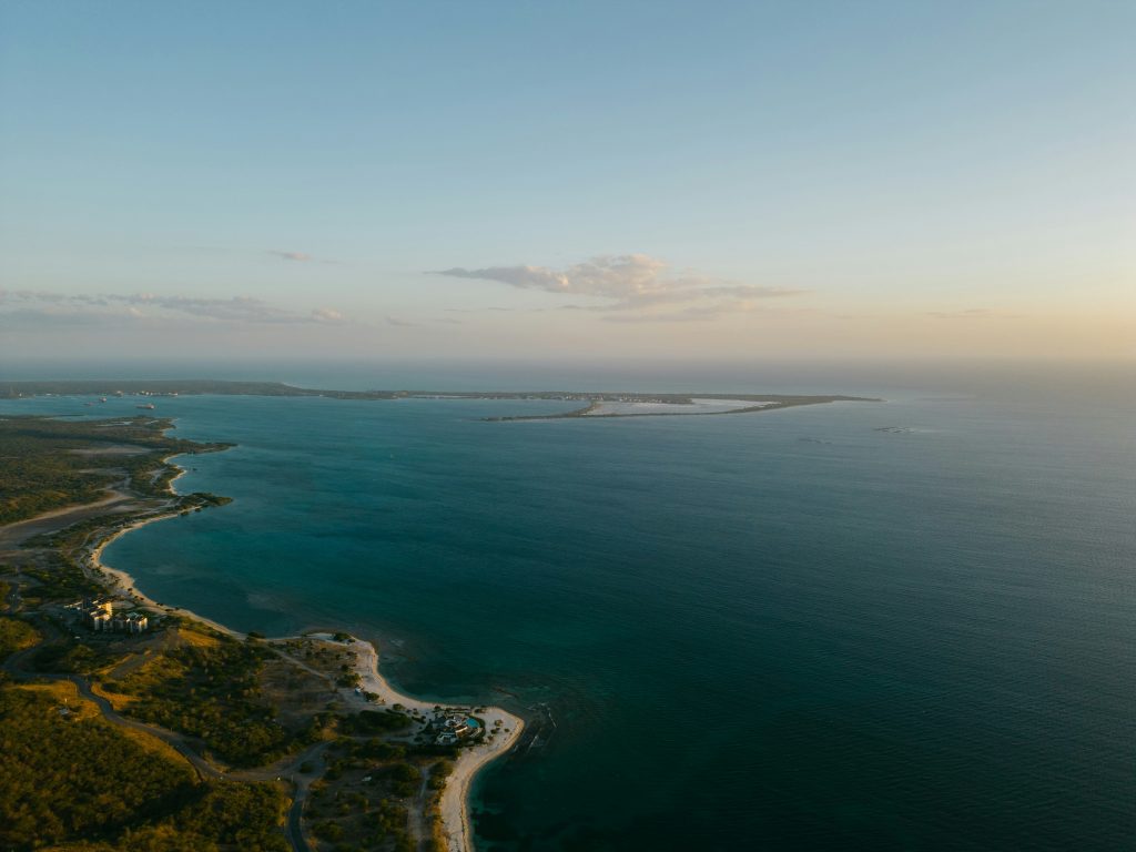 Aerial view of a coastline with clear blue-green water, sandy beaches, and patches of green vegetation, taken at sunset with soft, golden light and a partly cloudy sky.