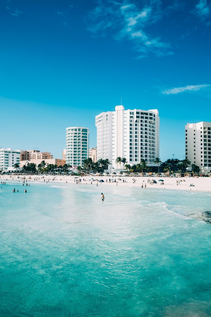 White high-rise hotels and palm trees line a busy sandy beach as people swim and relax near turquoise ocean water under a bright blue sky.