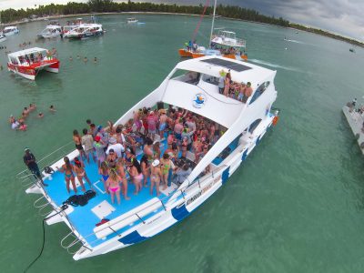 A large white boat crowded with people in swimwear floats on a greenish-blue ocean. Several other boats are nearby, and many people are swimming in the water. The background shows a cloudy sky and a distant treeline.